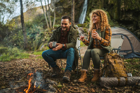 Couple camping in the forest near a campfire, with backpack and night vision goggles prepared for outdoor adventure, tent and waterfall in the background