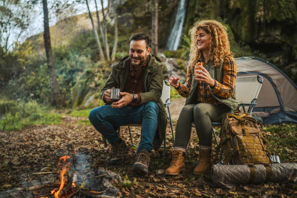 Couple camping in the forest near a campfire, with backpack and night vision goggles prepared for outdoor adventure, tent and waterfall in the background